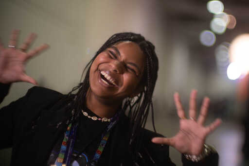 A woman with long braids laughs joyfully with both hands raised and fingers spread wide, wearing a black jacket and colorful lanyard.