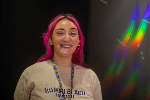 A woman with bright pink hair smiles warmly, wearing a beige 'Waikiki Beach Hawaii' sweatshirt and colorful lanyard, with rainbow light streaks in the background.