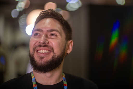 A bearded man smiles and gazes upward, wearing a black shirt and colorful lanyard, with bokeh lights in the background.