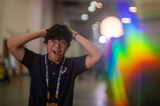 A young man with curly hair and glasses holds both hands on his head in a surprised or excited expression, wearing a dark polo shirt and colorful lanyard, with vivid rainbow bokeh behind him.