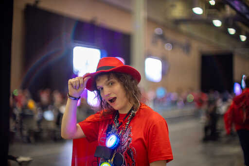 Aask, in a red Goon shirt, red fedora like the Red Hat from Linux, and a red cape, doffing her hat and smiling with mouth open, with an empty talking track behind her
