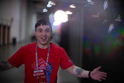A man in a red graphic t-shirt grins with one arm outstretched, showing a tattoo on his forearm, with prismatic light effects visible behind him.