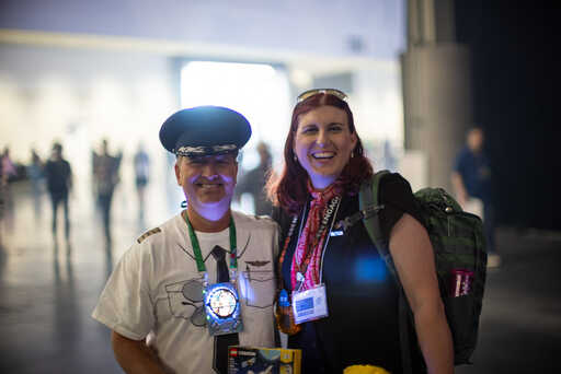 Spanky in a pilot-themed T-shirt and Zap dressed like a stewardess, from Aerospace Village, smiling in front of a dark hallway with the light of day shining past blurry silhouettes